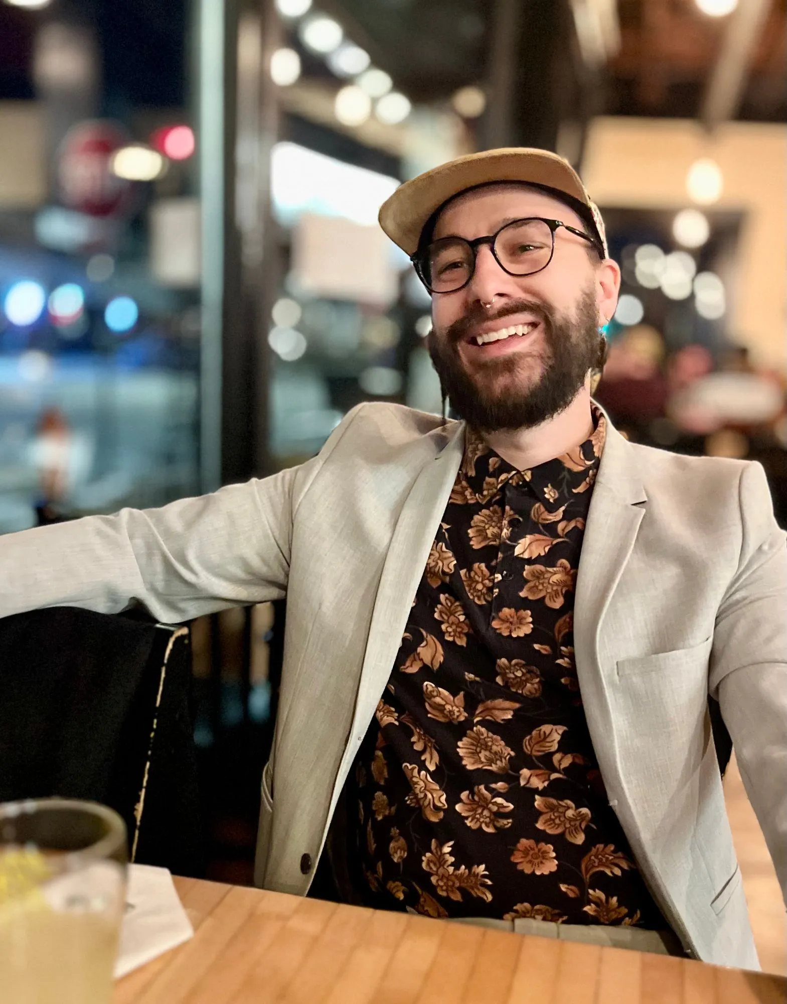 Michael VanDyke laughing at a restaurant table at night, in a tan cap, round glasses, a light gray blazer, and a black shirt with a copper floral print.